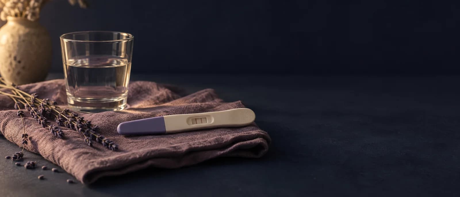 A positive home pregnancy test on a folded linen cloth, beside a glass of water and a sprig of dried lavender