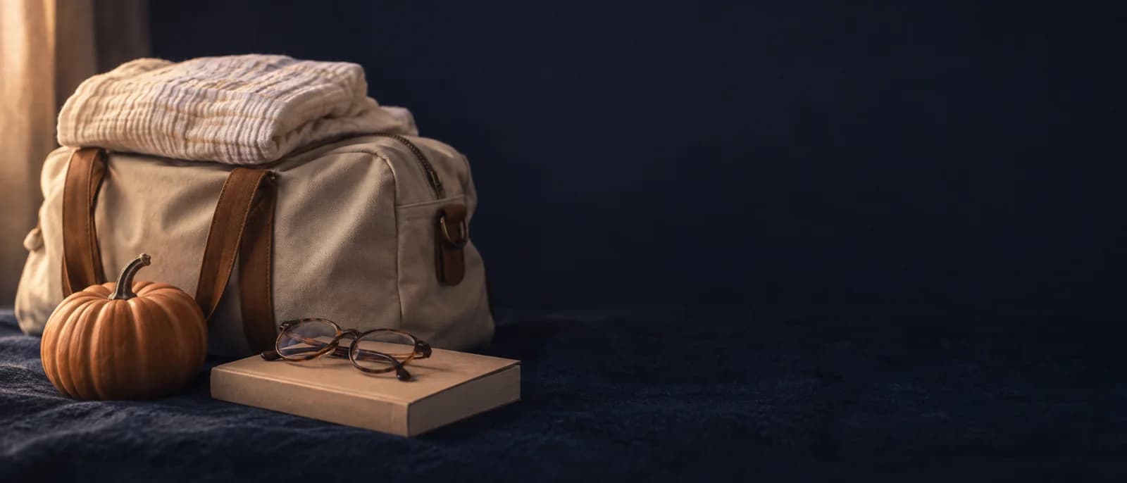 A packed hospital bag with a folded muslin blanket on top, a small pumpkin and reading glasses on a book